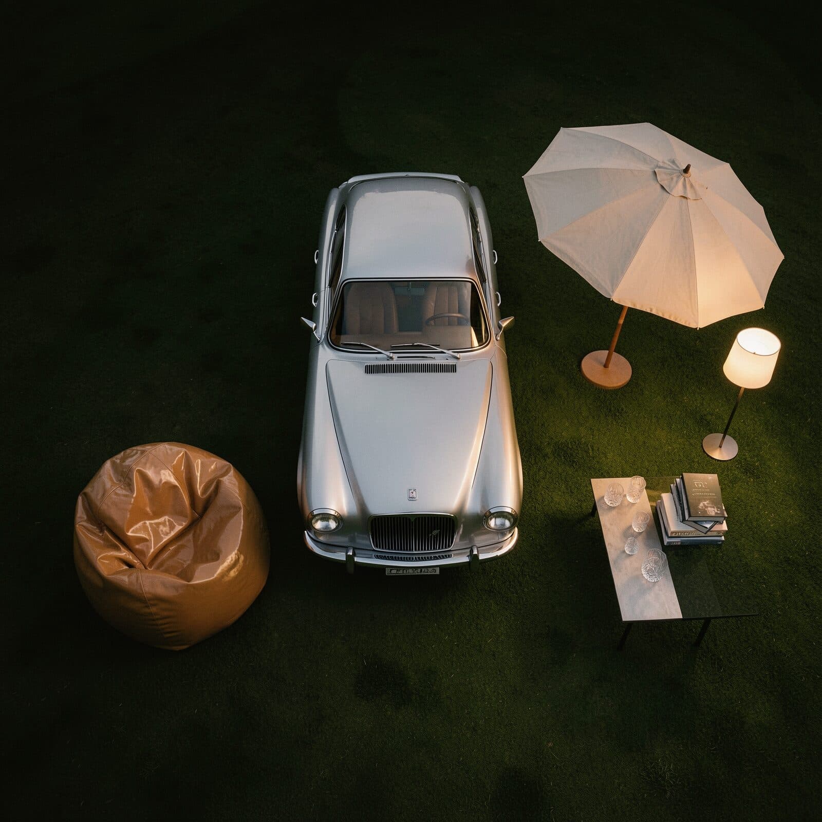 Overhead view at night of a silver saloon car parked on dark grass, a tan leather beanbag to its left, and to its right a parasol, a lit floor lamp and a low table set with glasses and stacked books.