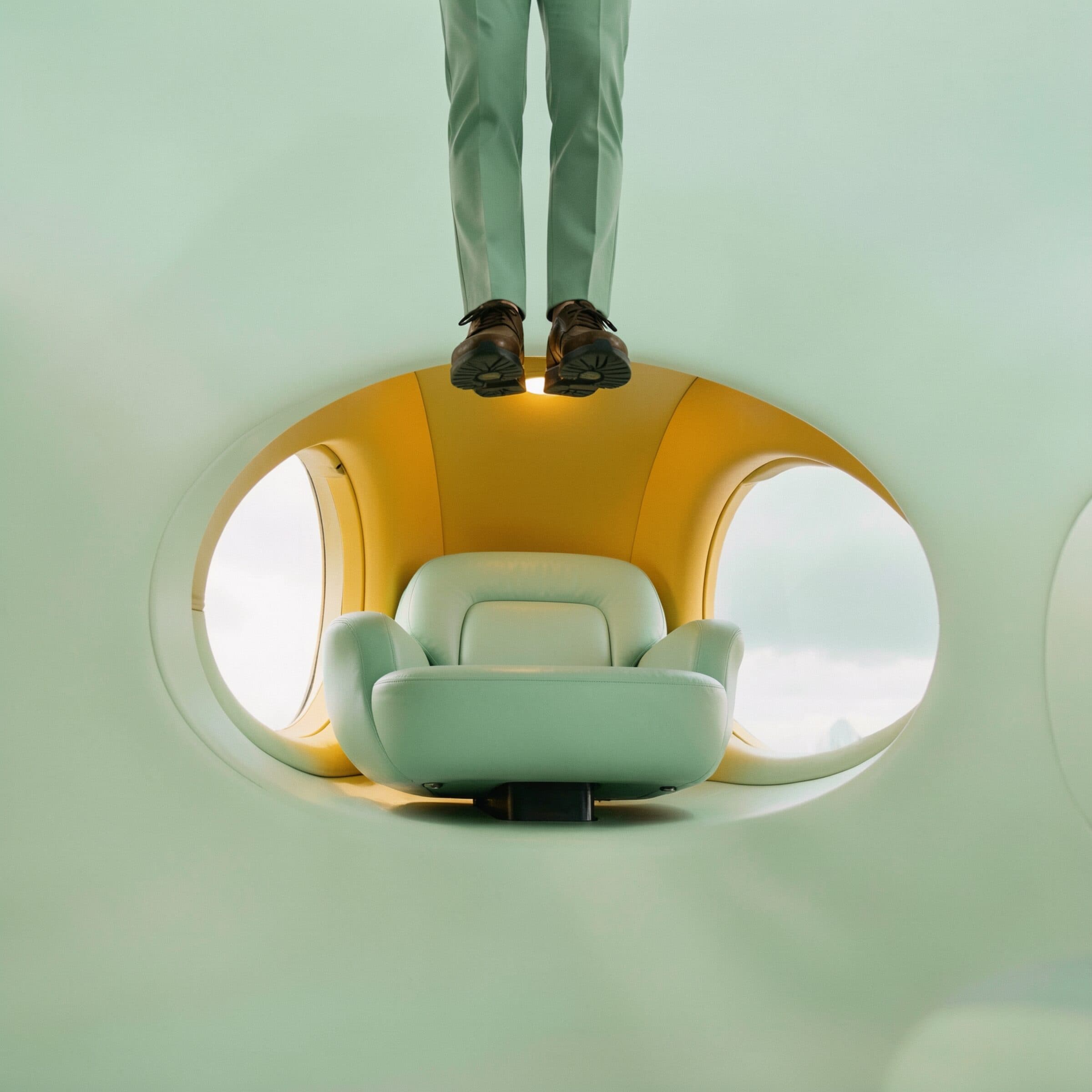 A pair of legs in mint-green trousers and brown brogues stands on the outside of a rounded cabin shell, mint-green above and below. Inside the opening, lit yellow, sits a mint leather armchair with cloud beyond the two oval windows either side of it.
